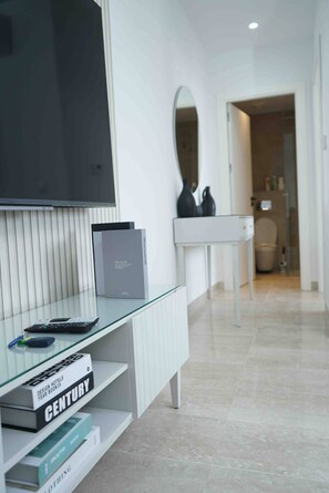 Una habitación de hotel moderna con una televisión de pantalla plana montada en la pared, una mesa consola de vidrio con libros, y un baño visible a través de una puerta abierta.