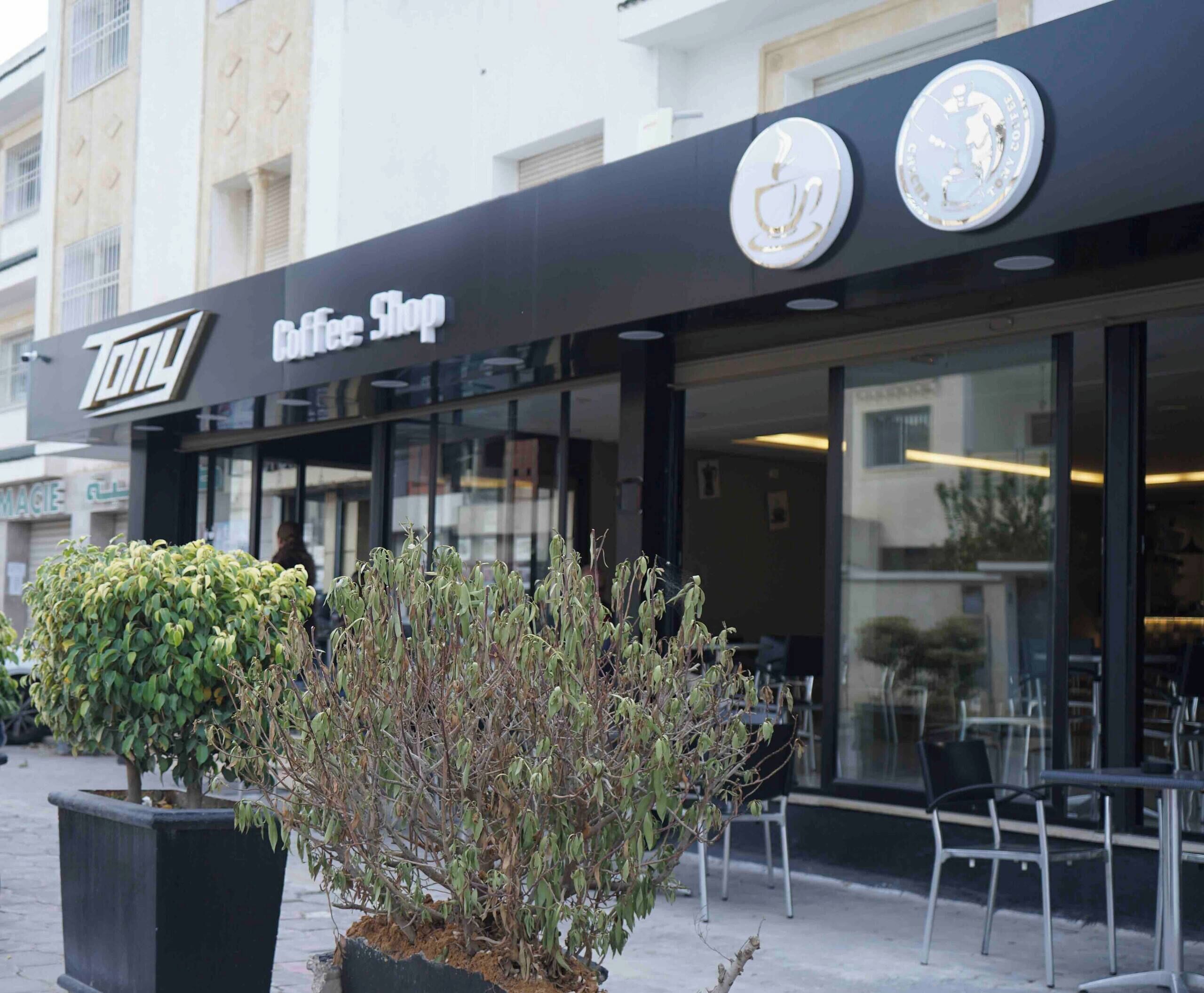 Een coffeeshop met een zwart luifel en een bord waarop 'Coffee Shop' staat. Buiten tafels en stoelen zijn zichtbaar.