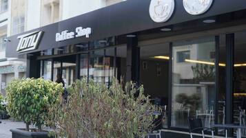Een coffeeshop met een zwart luifel en een bord waarop 'Coffee Shop' staat. Buiten tafels en stoelen zijn zichtbaar.