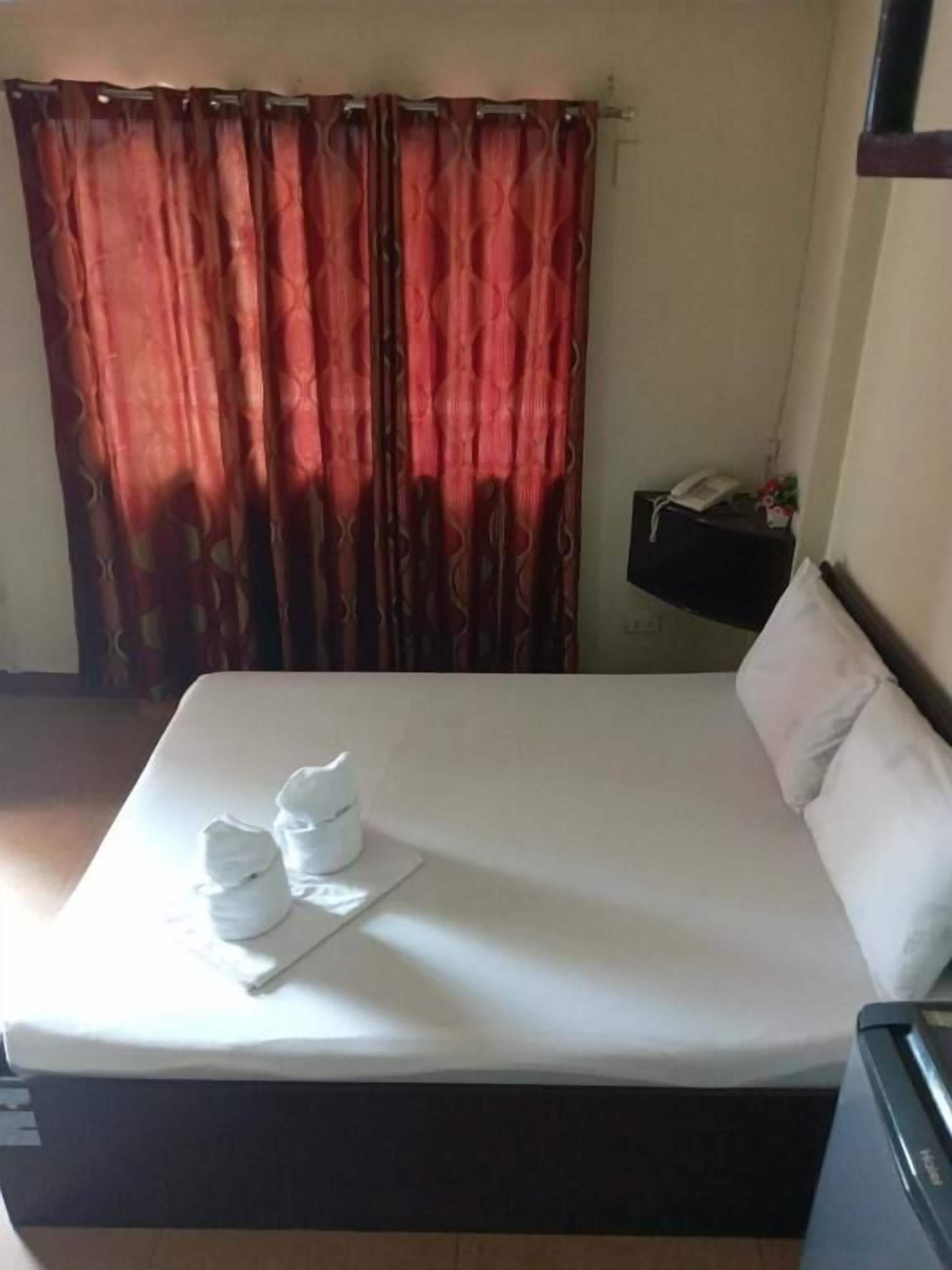 Una cama bien hecha con edredón blanco y almohadas, una mesita de noche con un teléfono y un objeto pequeño, y una ventana con cortinas rojas.