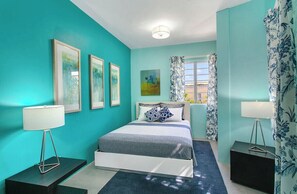 Un dormitorio con paredes color turquesa, una cama con estructura blanca, dos mesitas de noche con lámparas y cuadros en la pared.