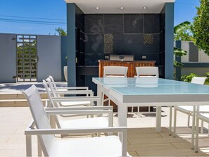 Un patio con una mesa de vidrio, sillas blancas, un mostrador de madera y una parrilla.