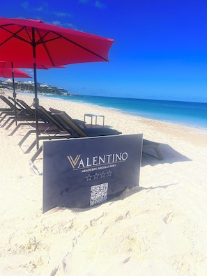 Schild am Strand für das Valentino Resort mit einem QR-Code, einem roten Sonnenschirm und Liegestühlen.