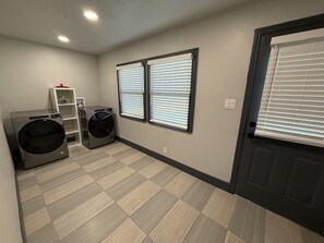 A laundry room with two washing machines, a shelf with a container, and two windows with blinds.