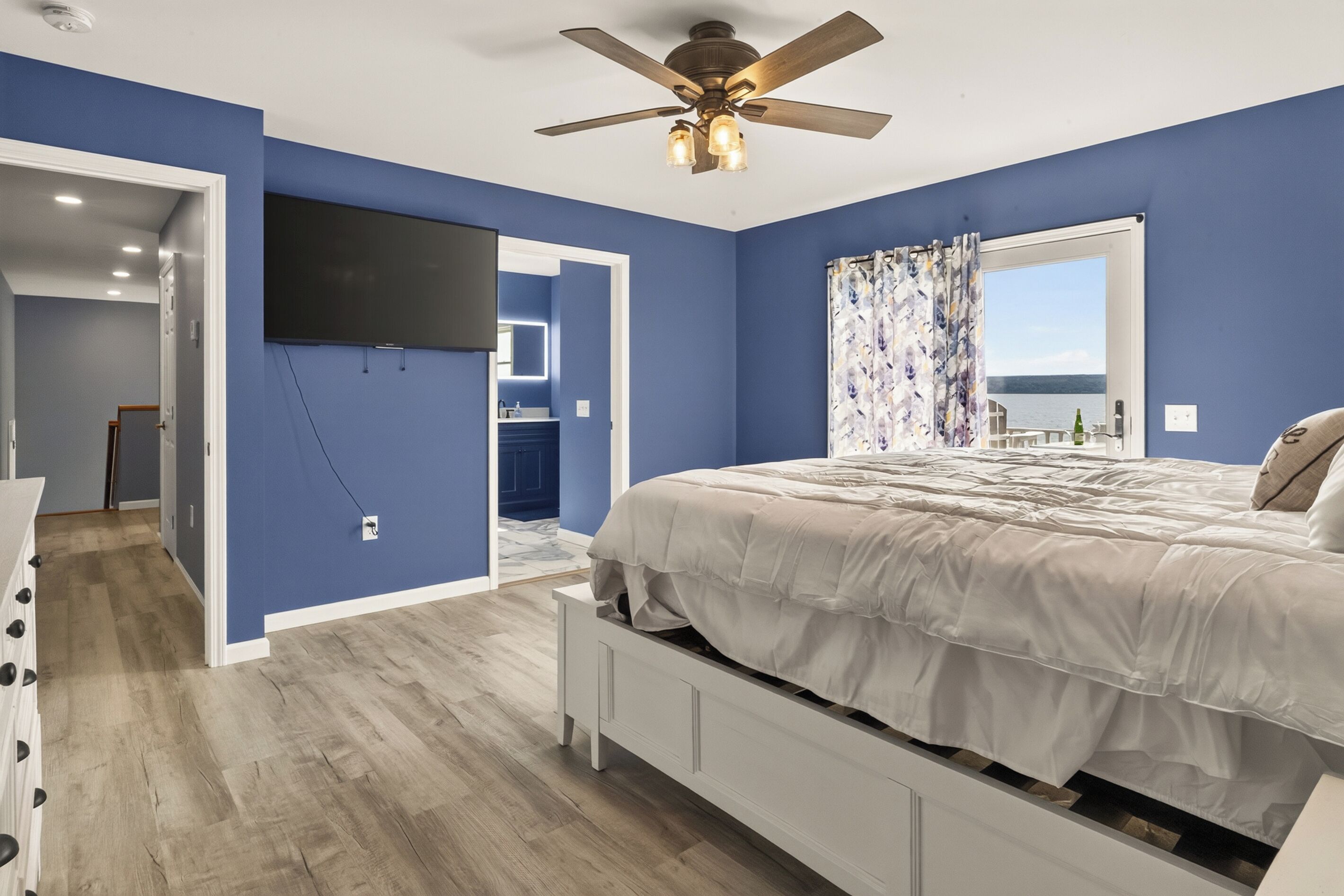 Une chambre à coucher avec un lit, un ventilateur de plafond, une télévision et une vue sur la mer par une fenêtre.