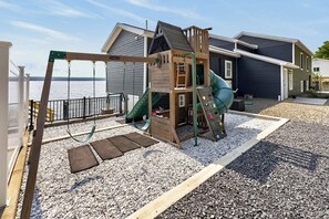 Un parque infantil con columpios, tobogán y estructura para escalar, junto a una casa con vista al agua.