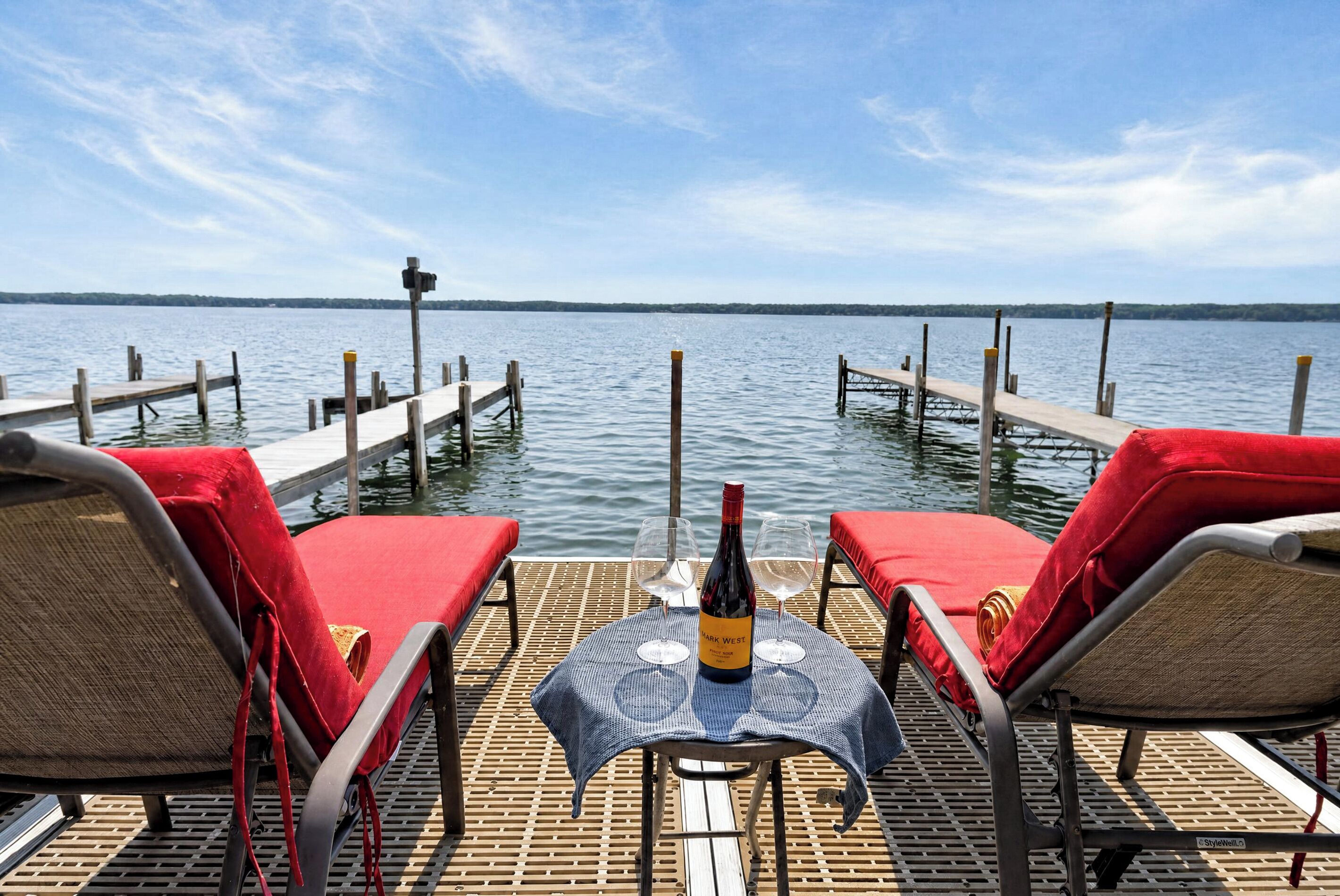Une terrasse avec deux chaises longues rouges, une table avec une bouteille et des verres, et une vue sur un lac paisible avec un quai en arrière-plan.