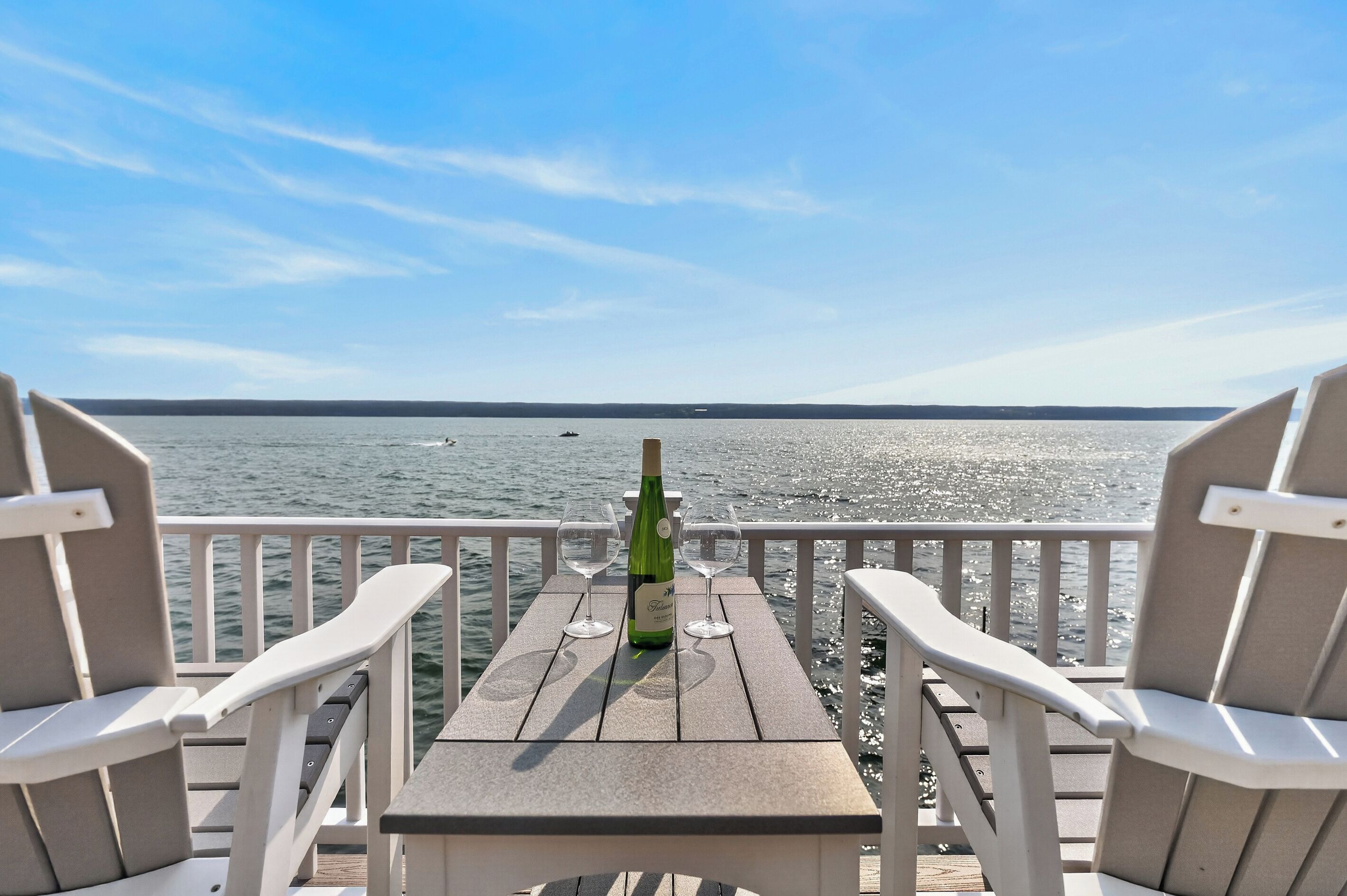Een houten terras met twee witte stoelen en een tafel, met uitzicht op een kalm wateroppervlak en een heldere lucht.