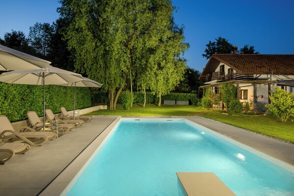 A swimming pool with lounge chairs and an umbrella, surrounded by greenery and a house.