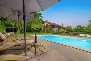 A swimming pool with lounge chairs and an umbrella, surrounded by greenery and a house.