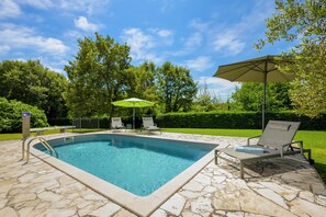 Piscina rectangular con baldosas de piedra, rodeada de sillas de descanso y sombrillas, con árboles y un cielo despejado de fondo.