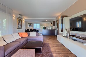 Ein modernes Wohnzimmer mit Sofa, Hocker und Couchtisch. Im Hintergrund ist eine Küche mit Essbereich zu sehen.