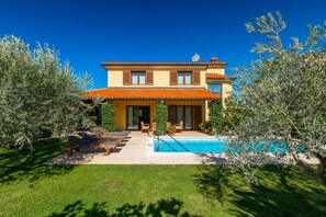 Ein zweistöckiges Haus mit Pool, Sitzgelegenheiten im Freien und Olivenbäumen.