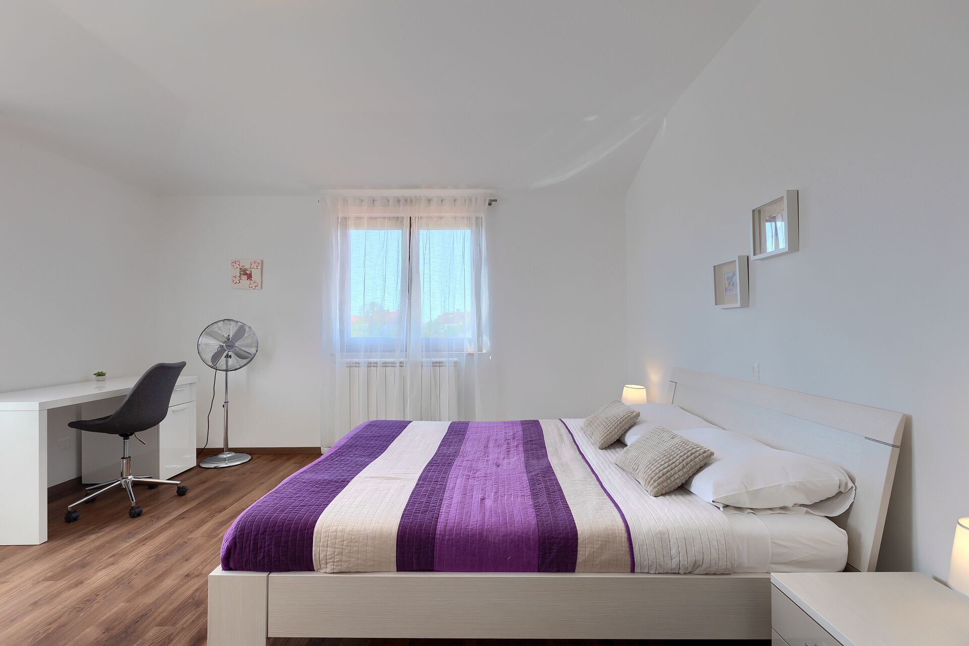 Ein Schlafzimmer mit einem Bett, einem Schreibtisch, einem Stuhl, einem Standventilator und einem Fenster mit durchsichtigen Vorhängen.