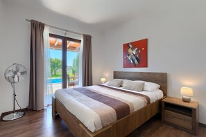 Ein Schlafzimmer mit einem hölzernen Bett, einem Standventilator, einem Nachttisch mit Lampe, einem Bild an der Wand und Blick auf einen Swimmingpool durch das Fenster.