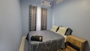 A bedroom with a bed, curtains, an air conditioning unit, a bedside table, and a patterned rug. - New Listing-Bungalow Verde Unit Two (Castries)