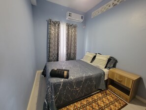 A bedroom with a bed, curtains, an air conditioning unit, a bedside table, and a patterned rug. - New Listing-Bungalow Verde Unit Two (Castries)