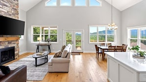 Un salón espacioso con chimenea, un sofá, una zona de comedor con mesa y sillas, y ventanas grandes que ofrecen vistas al exterior.