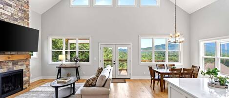 A spacious living room with a fireplace, a sofa, a dining area with a table and chairs, and large windows offering a view of the outdoors.