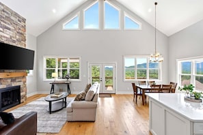 A spacious living room with a fireplace, a sofa, a dining area with a table and chairs, and large windows offering a view of the outdoors. - Highlander's Ridge | Sweeping Mountain Views 5 Min. from Black Mtn! (Black Mountain)