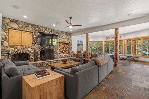 A spacious living area with a stone fireplace, comfortable seating, and large windows offering a view of the outdoors. - Luxury Mountain Retreat with Panoramic Views, Hot Tub, Game Room, & Endless Outdoor Living (Durango)