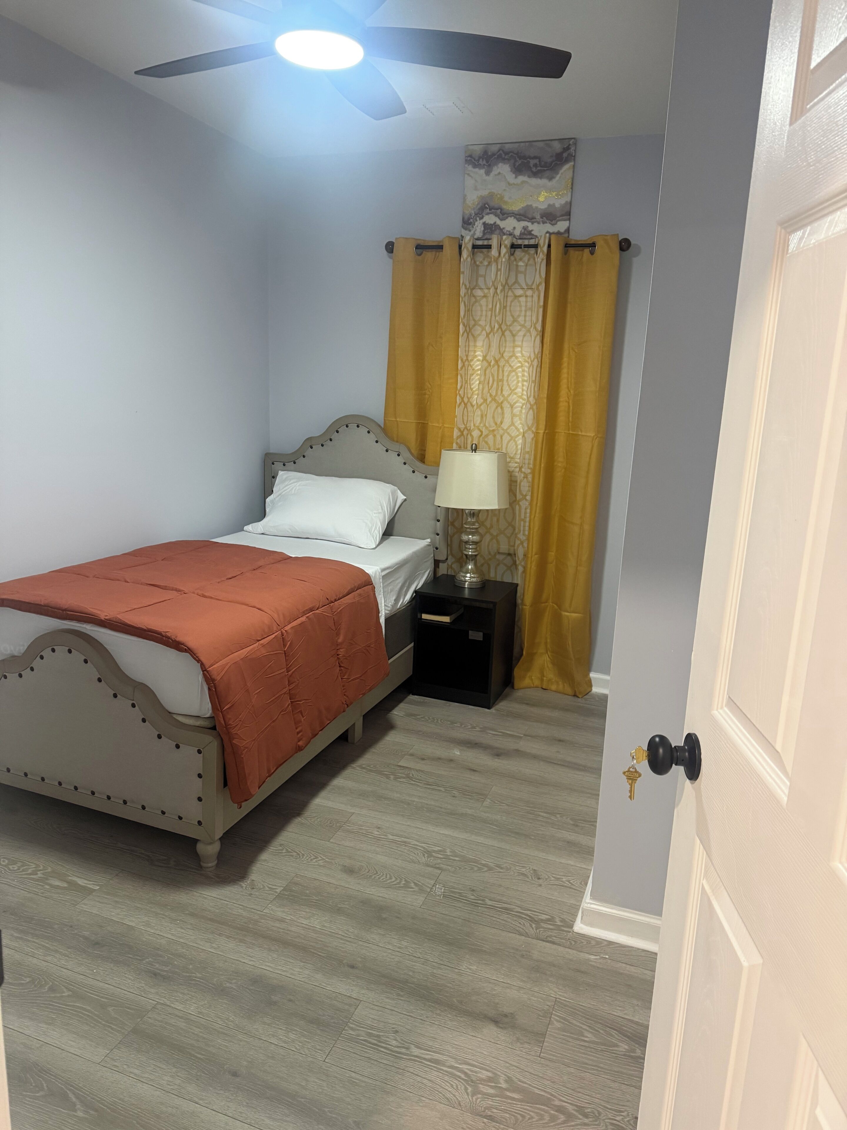 Un dormitorio con una cama, una mesita de noche, una lámpara, un ventilador de techo y una puerta.