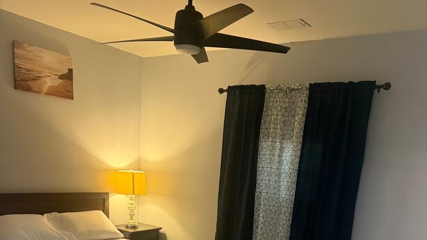 A bedroom with a bed, a nightstand, a lamp, a ceiling fan, and a window with curtains. - Spacious 5-Bedroom Home Near Atlanta Airport Area (Atlanta)