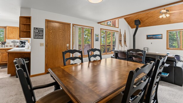 A dining area with a wooden table and chairs, a kitchen in the background, and large windows.