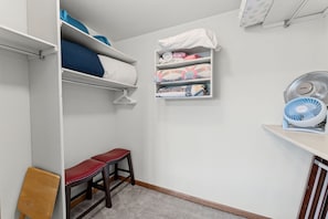 A bedroom with a bunk bed, a bedside table, a chair, a shelf with folded blankets, and a fan.