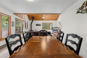 A dining room with a long wooden table and black chairs, a fireplace, and large windows with a view of trees.