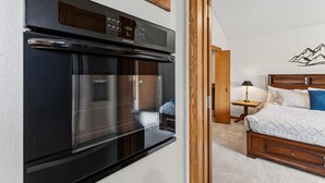 A kitchen with a modern black oven, a wooden cabinet above, and a bedroom with a bed, nightstand, and a wall-mounted lamp.
