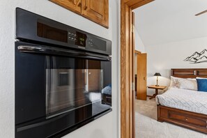 A kitchen with a modern black oven, a wooden cabinet above, and a bedroom with a bed, nightstand, and a wall-mounted lamp.