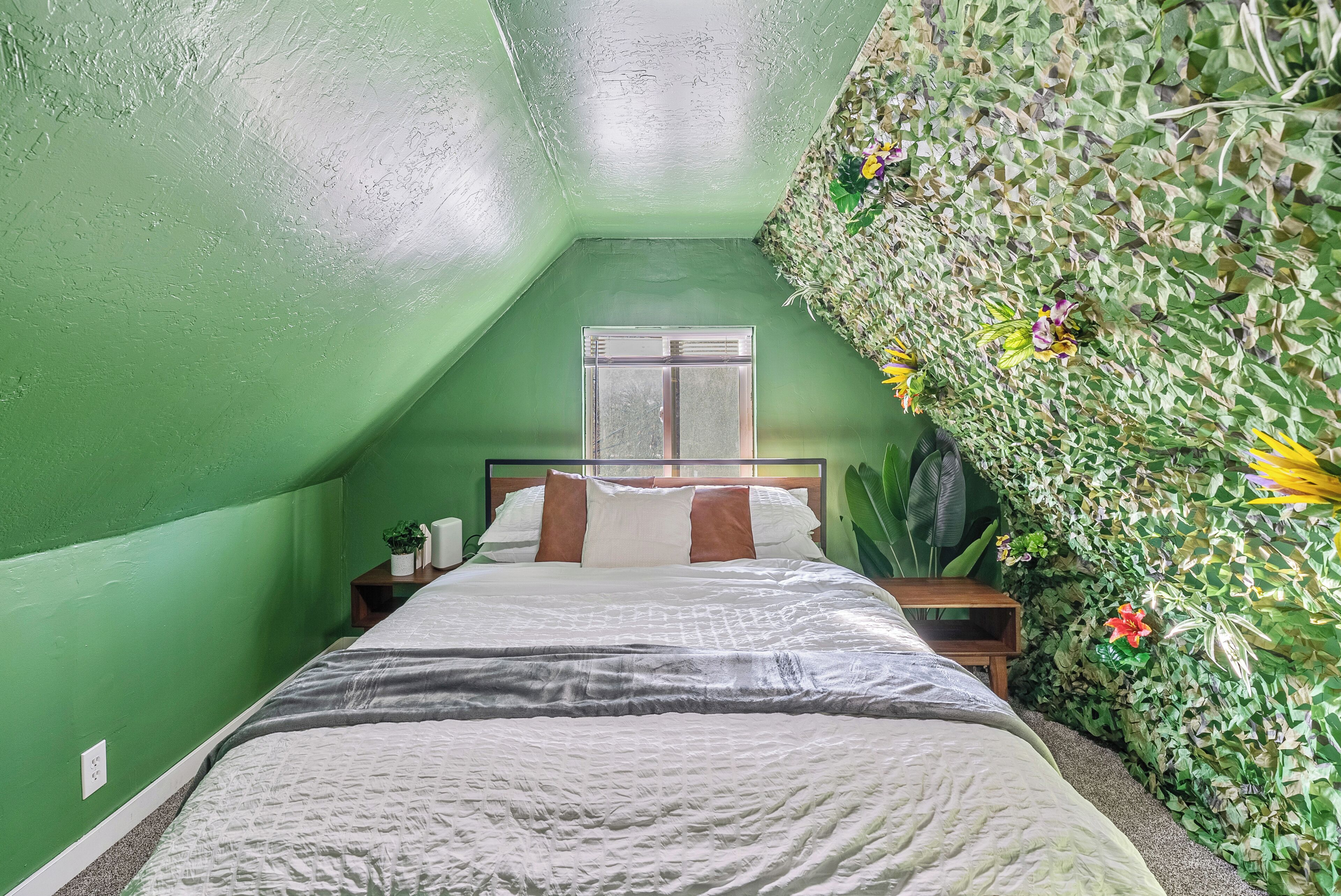 Ein Zimmer mit schräger Decke, einem Bett mit weißer Bettwäsche, einem Nachttisch mit Lampe und einer Wand mit grünem Blattmuster.