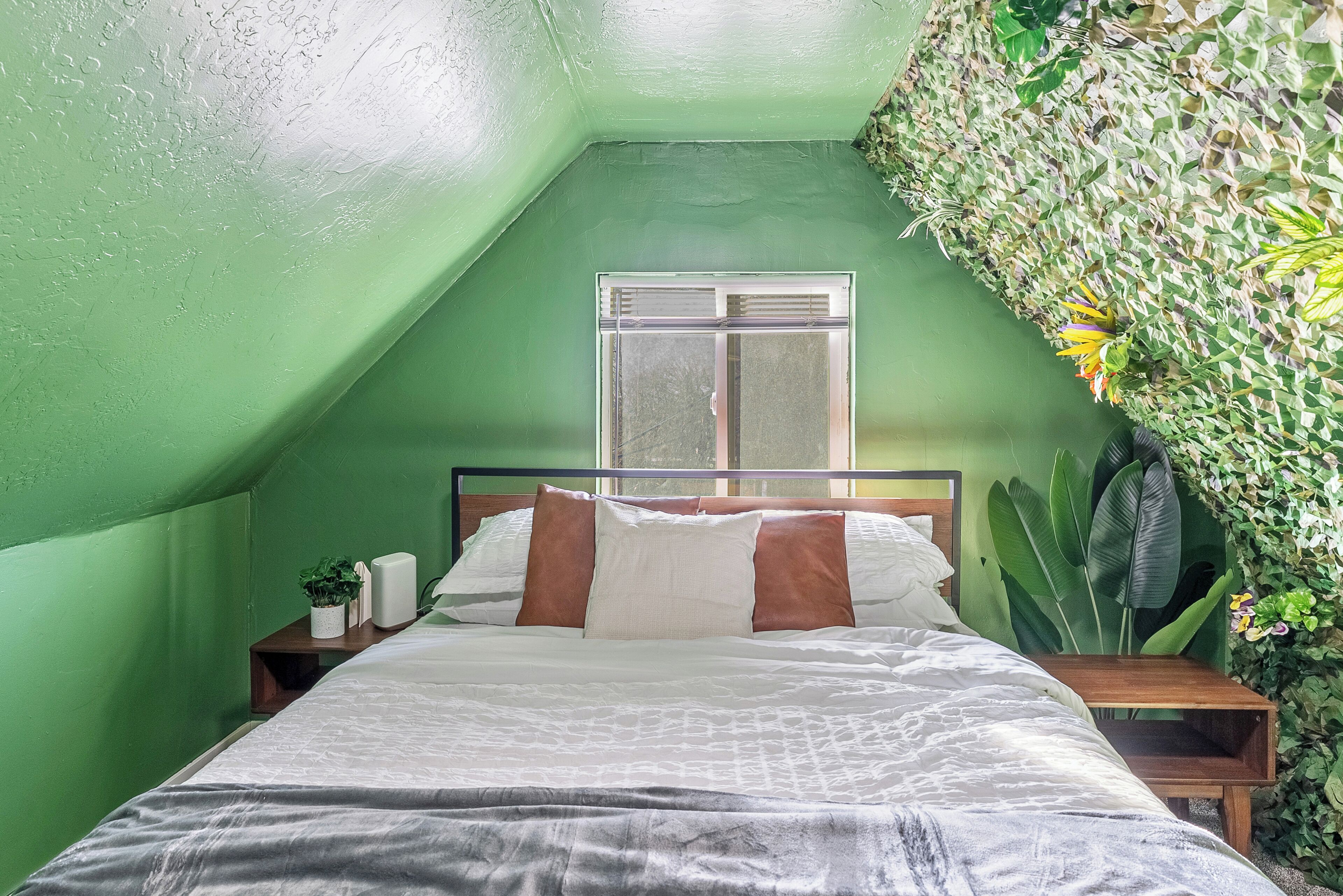 A bedroom with a sloped ceiling, a bed with white bedding and brown pillows, a bedside table with a lamp, and a window with a view of the outside.