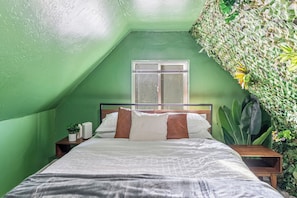 A bedroom with a sloped ceiling, a bed with white bedding and brown pillows, a bedside table with a lamp, and a window with a view of the outside. - Family-Friendly Provo Home w/ Parking (Provo)
