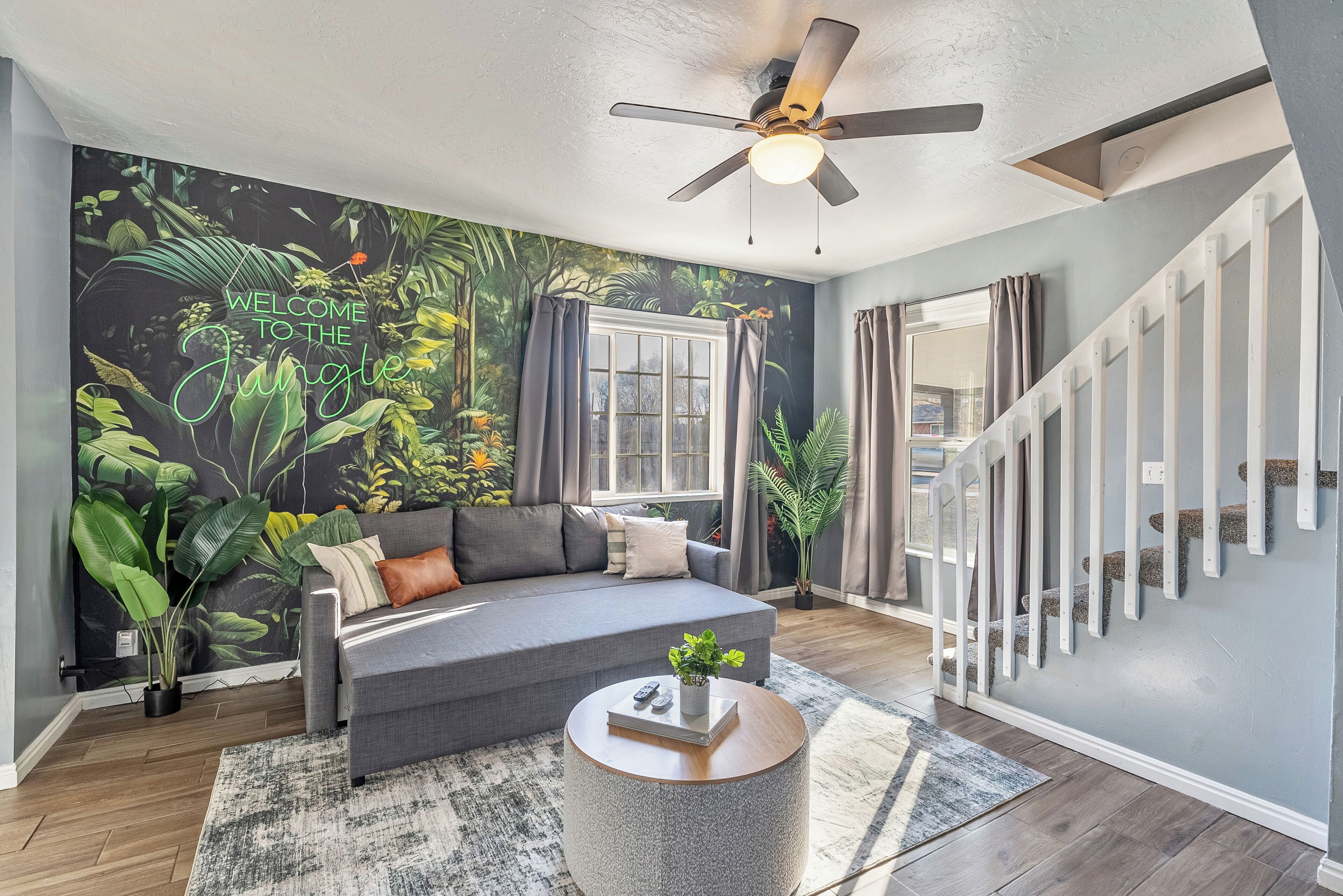 Un salon avec un canapé gris, une table basse ronde, un ventilateur de plafond, un escalier et une fresque murale sur le thème de la jungle.