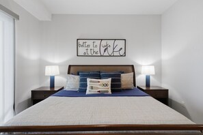 A neatly made bed with a blue bedspread and pillows, two bedside lamps, and a wall art piece. - The Golden Hour - Elevator & Main Channel Views! (Osage Beach)
