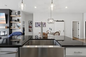 A modern kitchen with a black countertop, stainless steel appliances, pendant lighting, and a dining area with a white door. - The Golden Hour - Elevator & Main Channel Views! (Osage Beach)