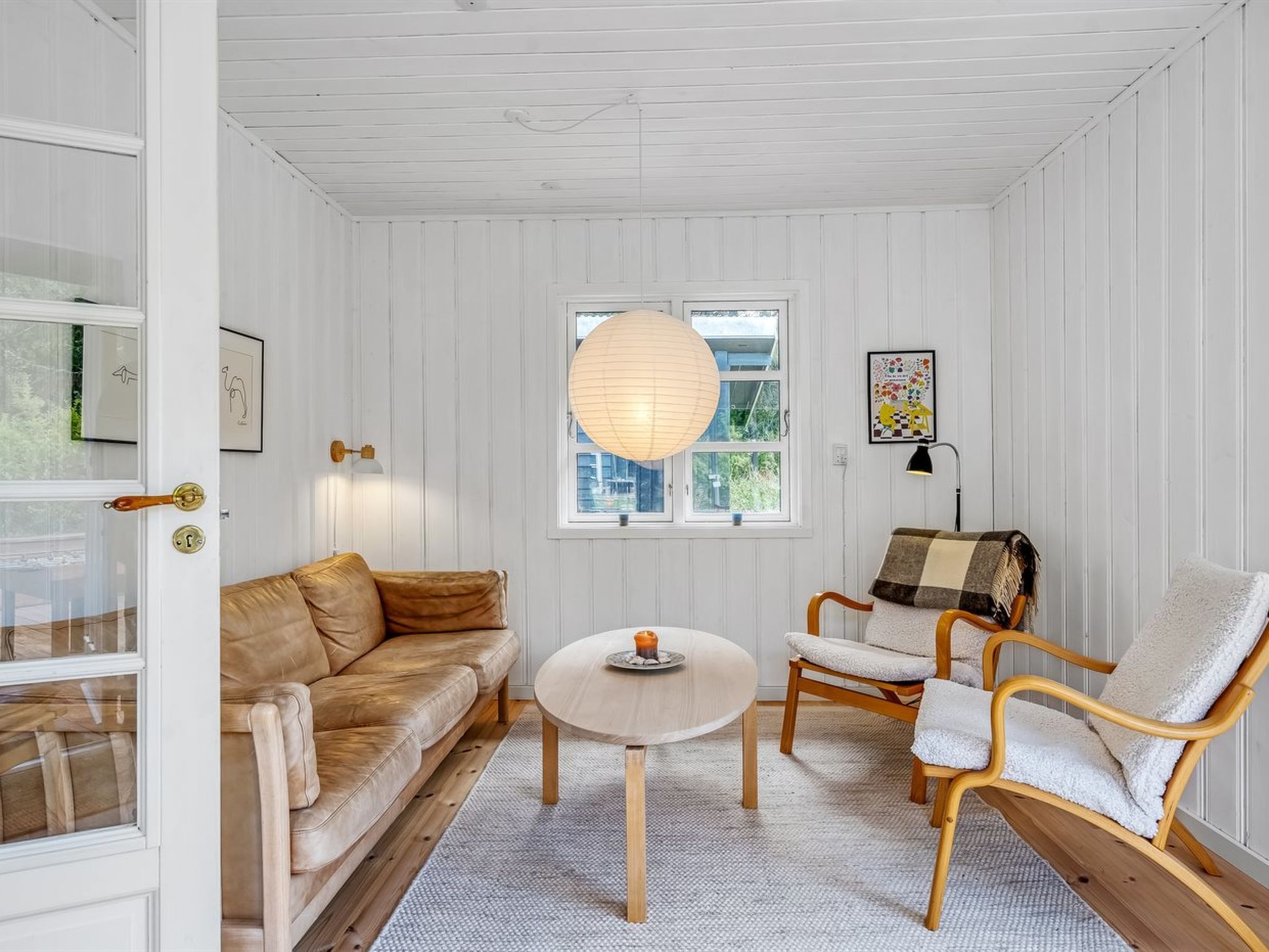 Une pièce avec un mur lambrissé blanc, un canapé en cuir marron, une chaise en bois recouverte d’une couverture, une table ronde avec une bougie et un luminaire suspendu.
