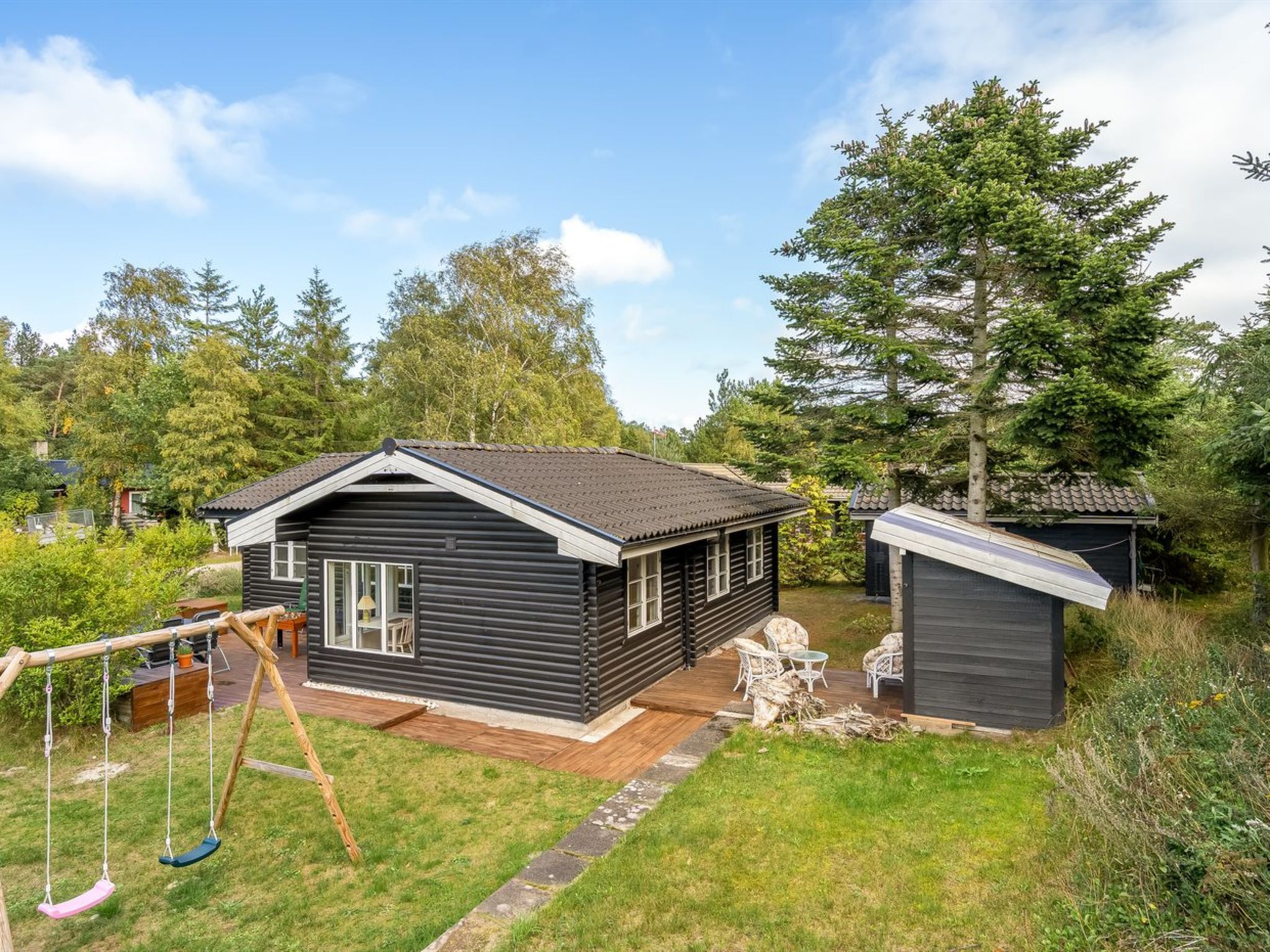 A wooden cabin with a porch and swing set, surrounded by trees and a small shed.