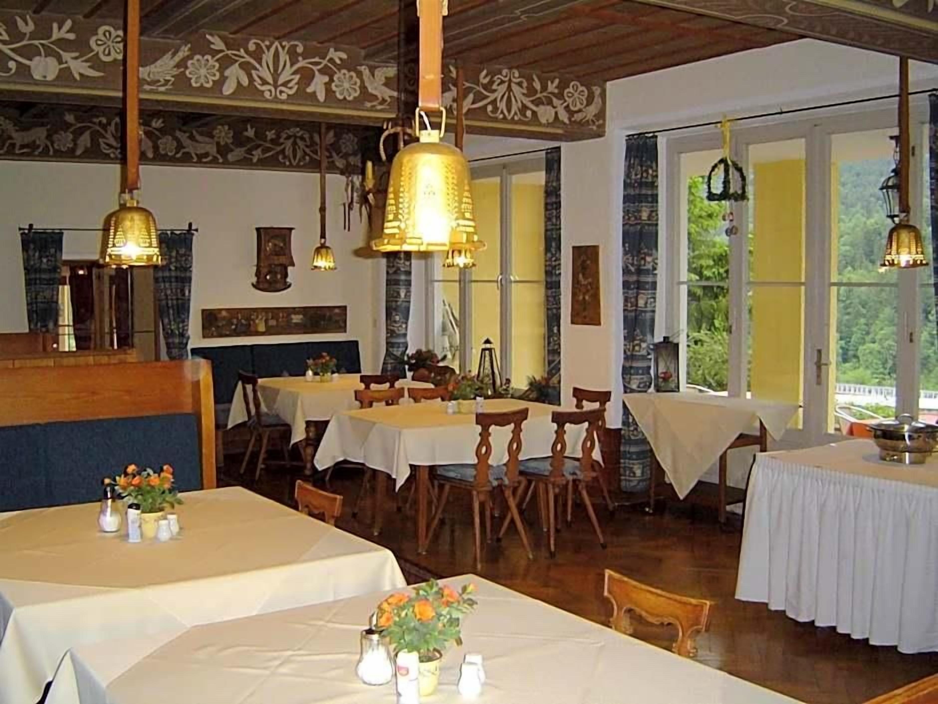 Ein Restaurant mit mehreren Tischen und Stühlen, Holzbalken an der Decke und großen Fenstern mit Vorhängen.