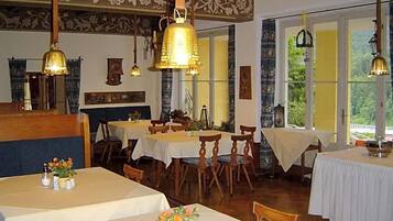 Ein Restaurant mit mehreren Tischen und Stühlen, Holzbalken an der Decke und großen Fenstern mit Vorhängen.