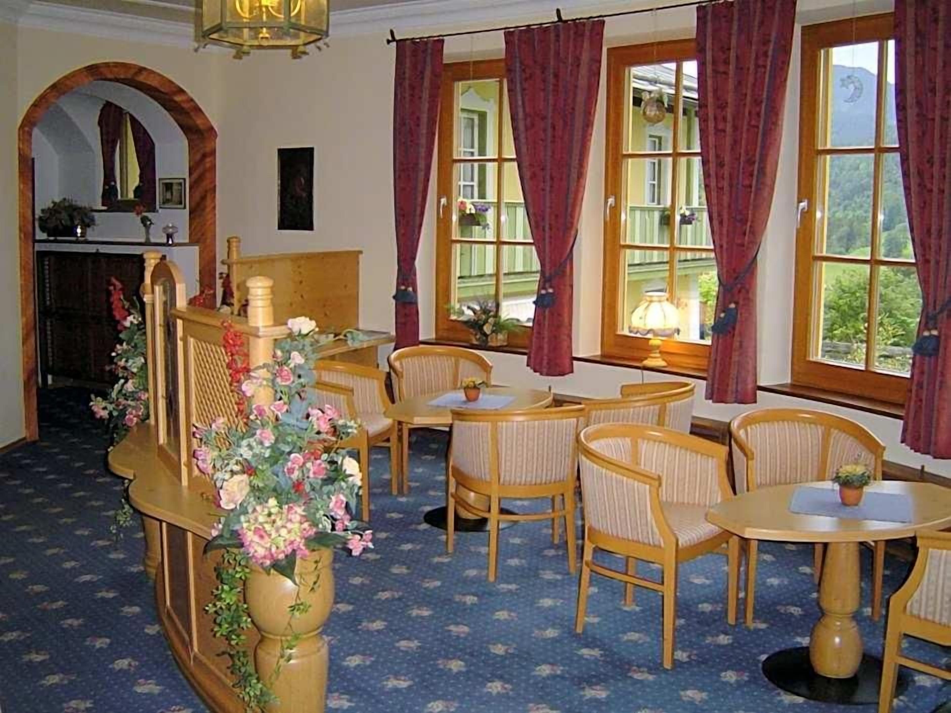 Ein Zimmer mit einem Holzbüro, mehreren Stühlen, einem kleinen Tisch und großen Fenstern mit Vorhängen.