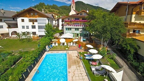 A swimming pool with lounge chairs and umbrellas, surrounded by houses and a church. - Wandergasthof Stern (Funes)