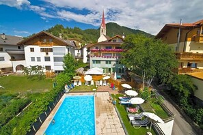A swimming pool with lounge chairs and umbrellas, surrounded by houses and a church. - Wandergasthof Stern (Funes)
