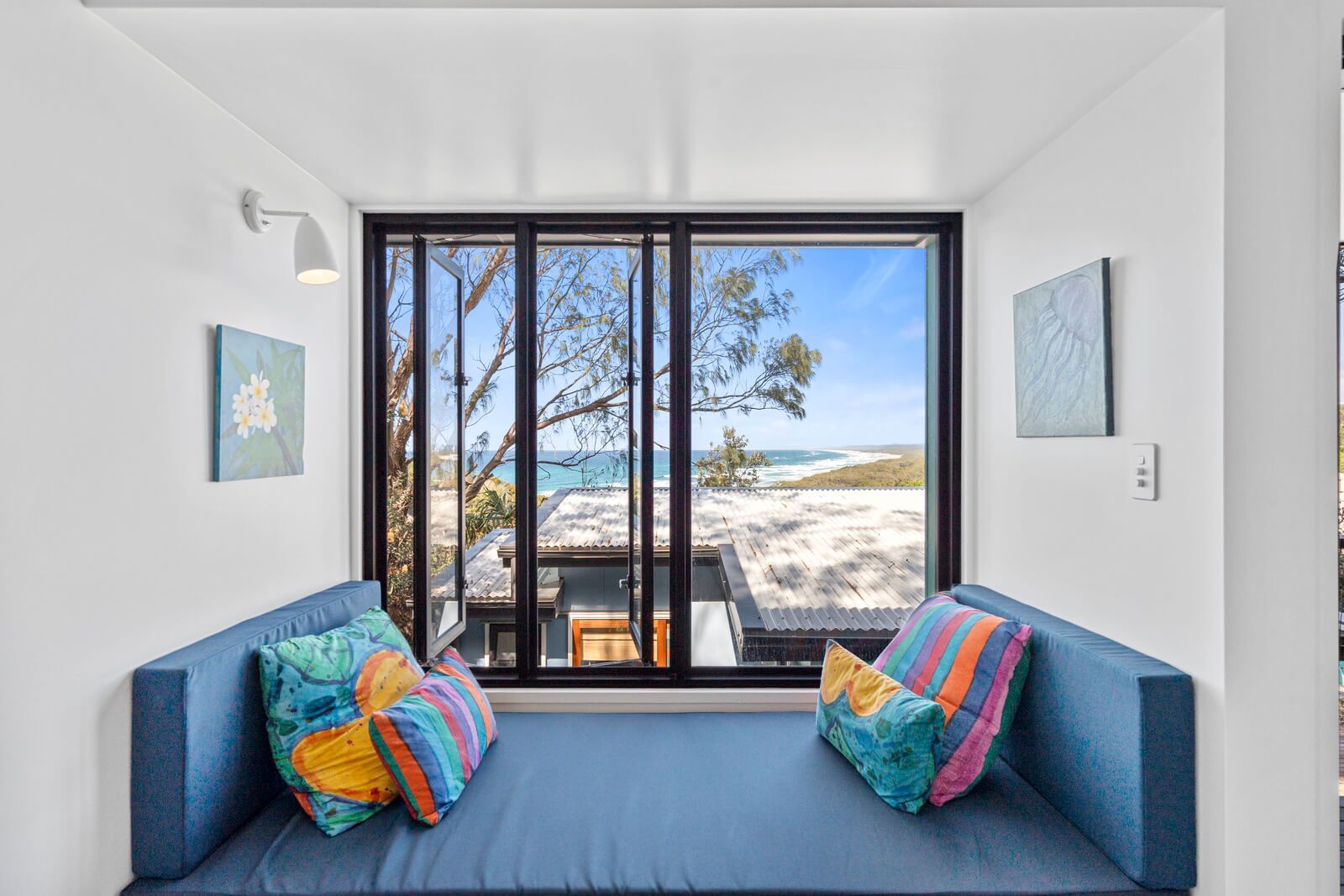 A room with a large window, two blue sofas with colorful pillows, and artwork on the walls.