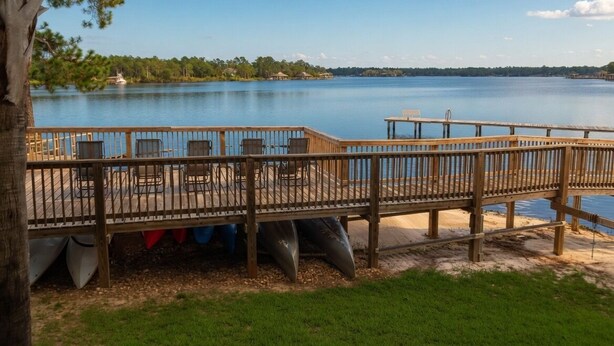 Una terraza de madera con sillas y mesas da a un lago tranquilo, rodeado de vegetación.