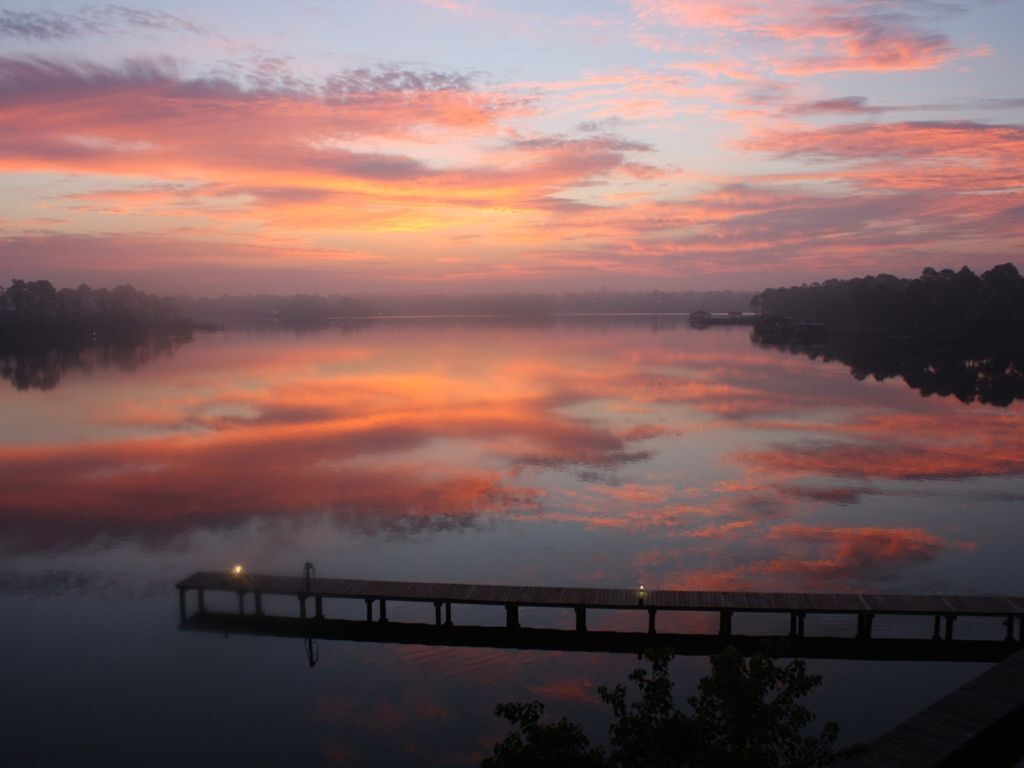 Une étendue d'eau calme reflétant le ciel au coucher du soleil, une jetée en bois s'étendant dans l'eau et une côte lointaine bordée d'arbres.