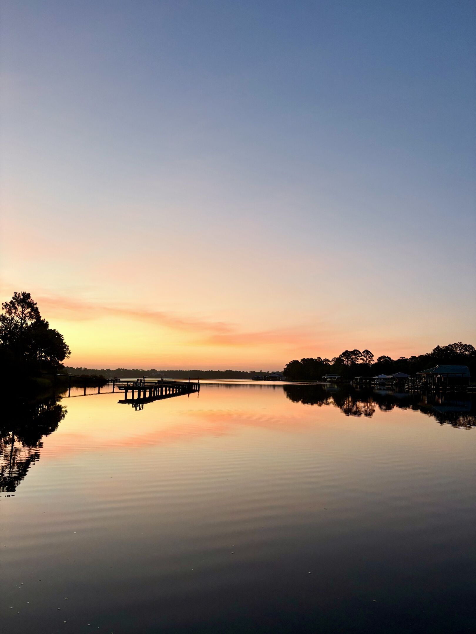 A calm lake with a pier extending into the water, surrounded by trees and buildings.