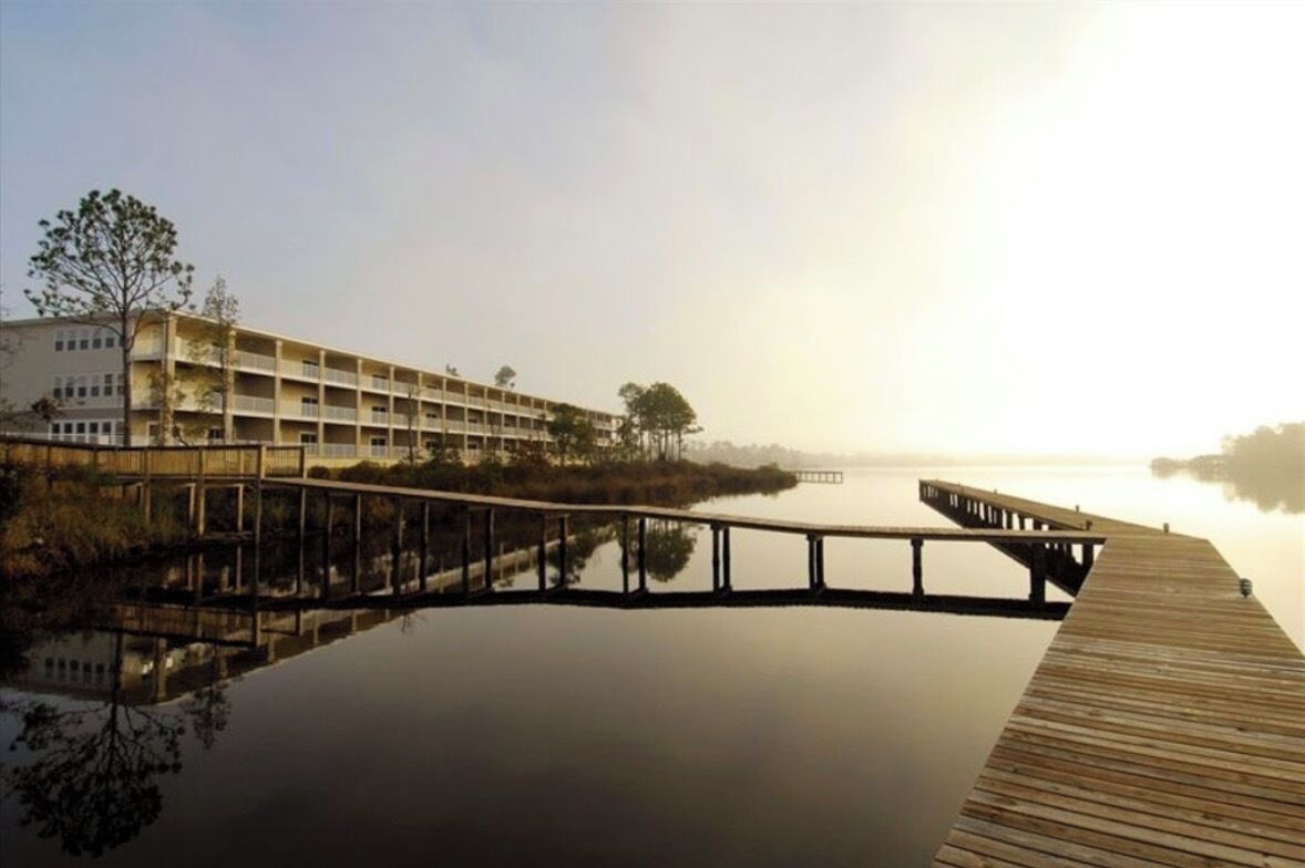 Un hôtel situé au bord de l’eau, doté d’une jetée en bois qui s’avance sur une étendue d’eau calme, entouré d’arbres et surplombé d’un ciel dégagé.